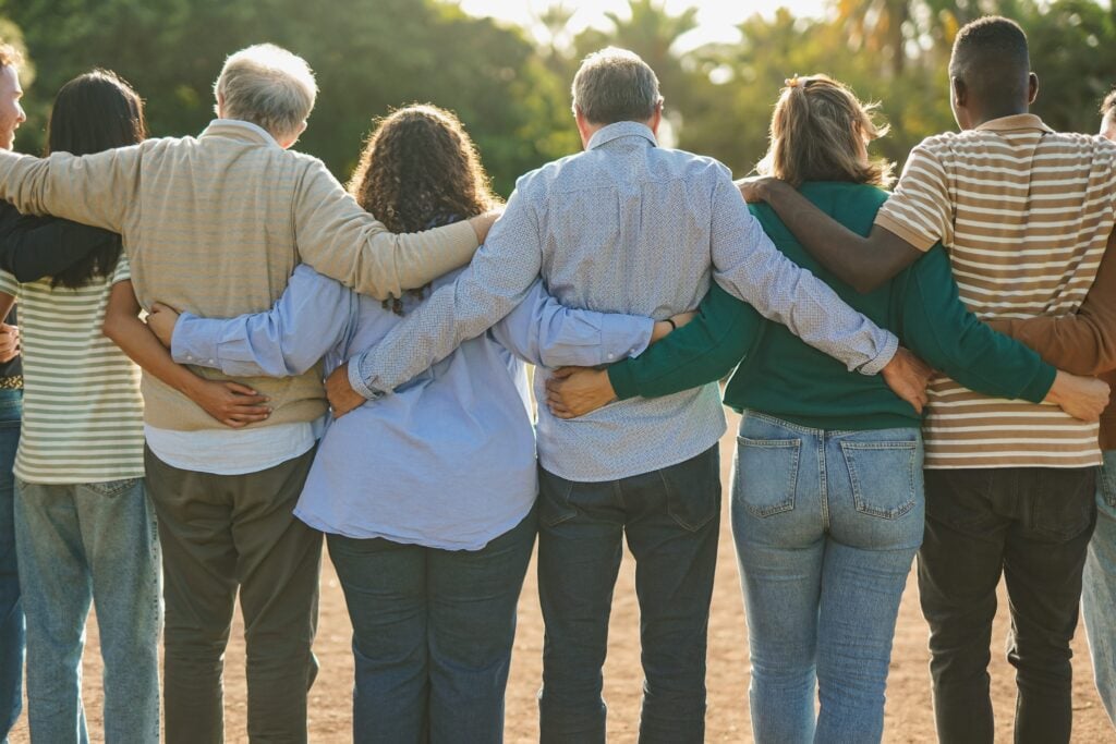 People seen from behind, standing outdoors in what appears to be a park area, with their arms wrapped around each other's shoulders in a show of unity and togetherness.
