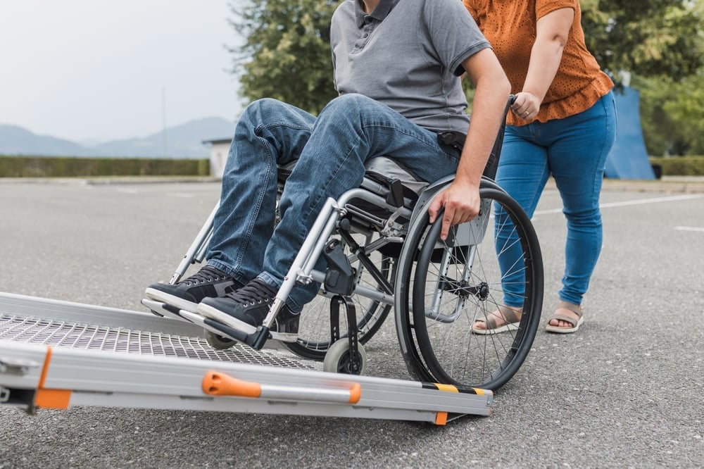 Person in a grey shirt and jeans is sitting in a manual wheelchair, being assisted by another person who is pushing them up a portable metal ramp.