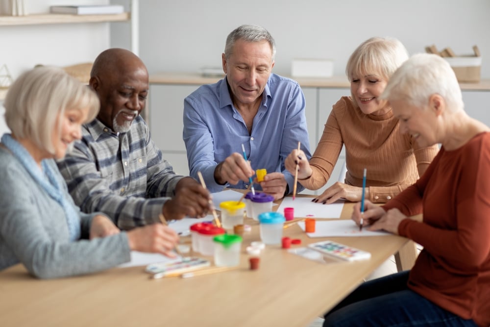 Five senior adults are seated around a light wooden table, engaging in an art activity.